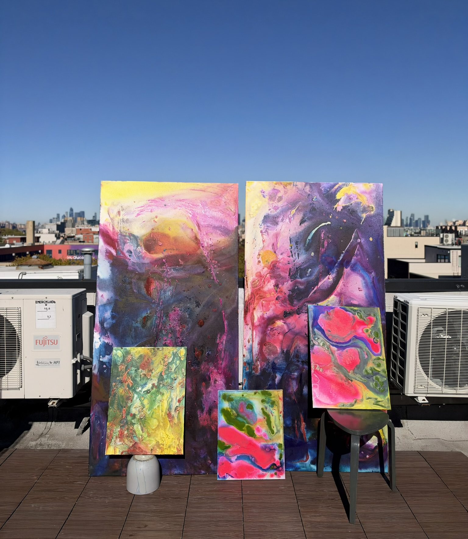 Five abstract paintings on a Brooklyn rooftop: two tall canvases in yellow, pink and violet with three small works in green, pink and blue propped in front, skyline behind.