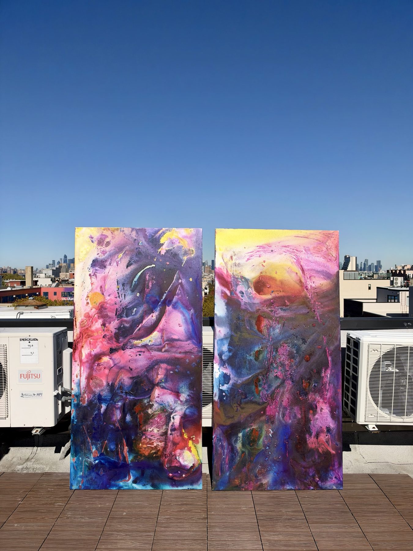 Two tall abstract paintings standing side by side on a rooftop, one dark purple and blue, one yellow and pink, with the Manhattan skyline behind.