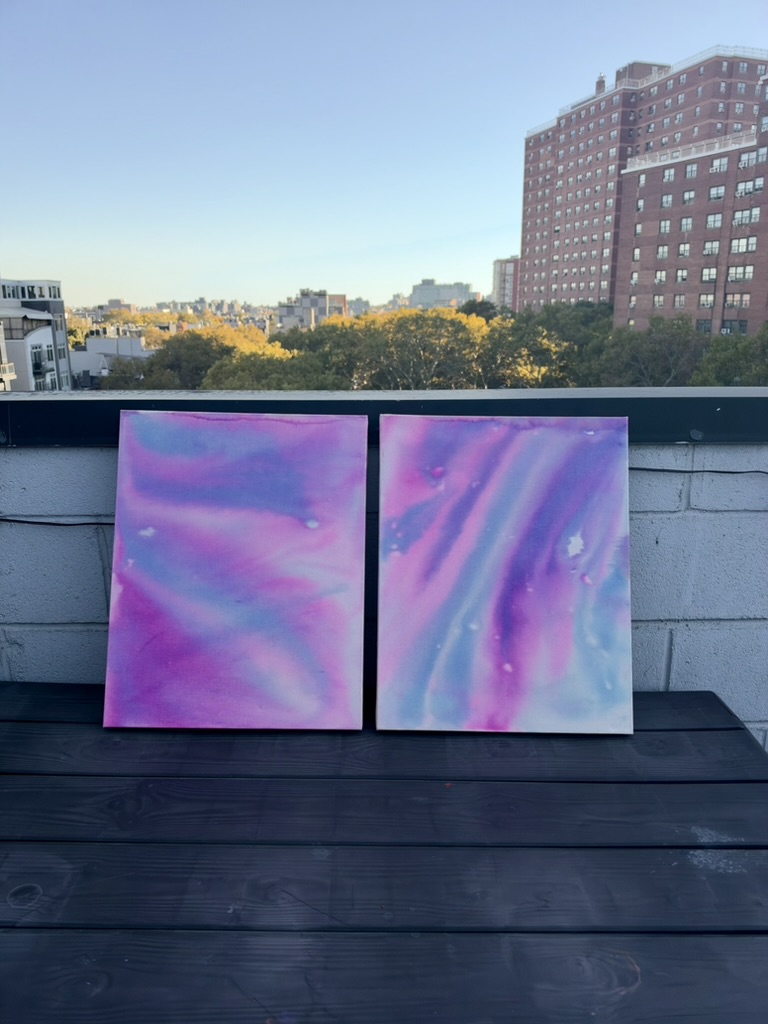 Two small canvases in soft pink, violet and blue washes standing side by side on a rooftop ledge at golden hour.