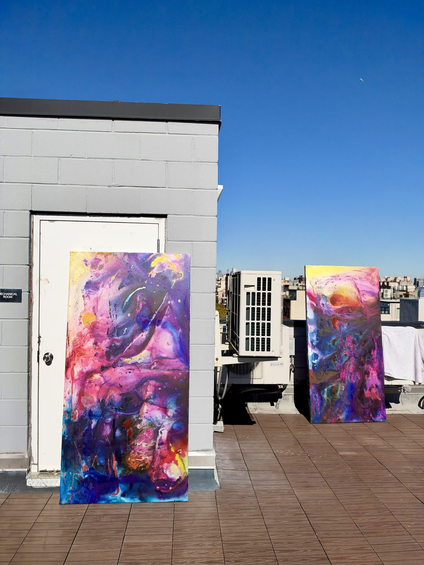 The two large paintings standing apart on a rooftop beside a grey block wall and a mechanical room door.