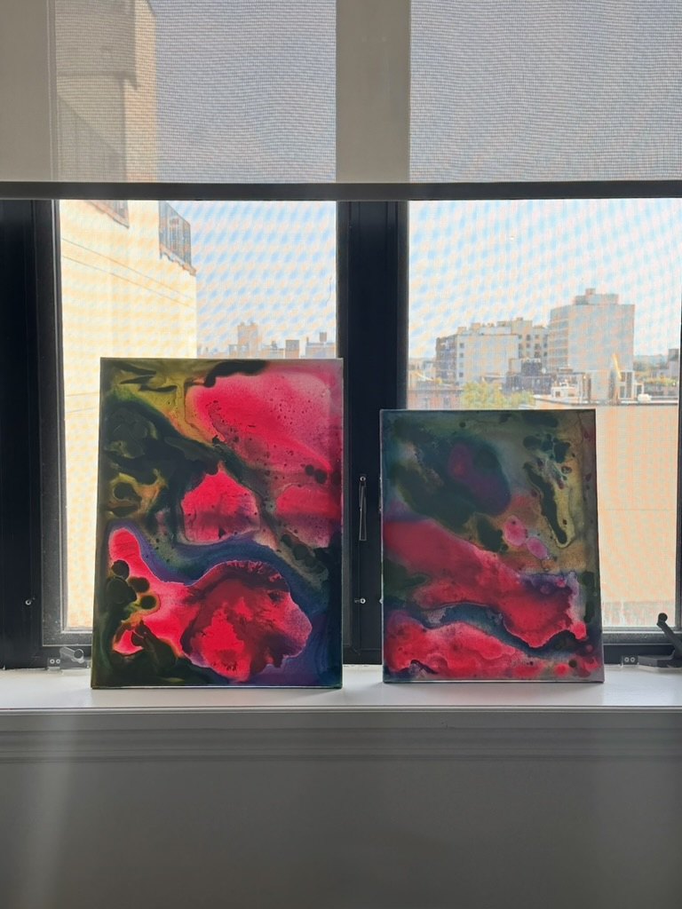 Two small canvases in hot pink, green and deep blue standing on a windowsill against the Brooklyn skyline.
