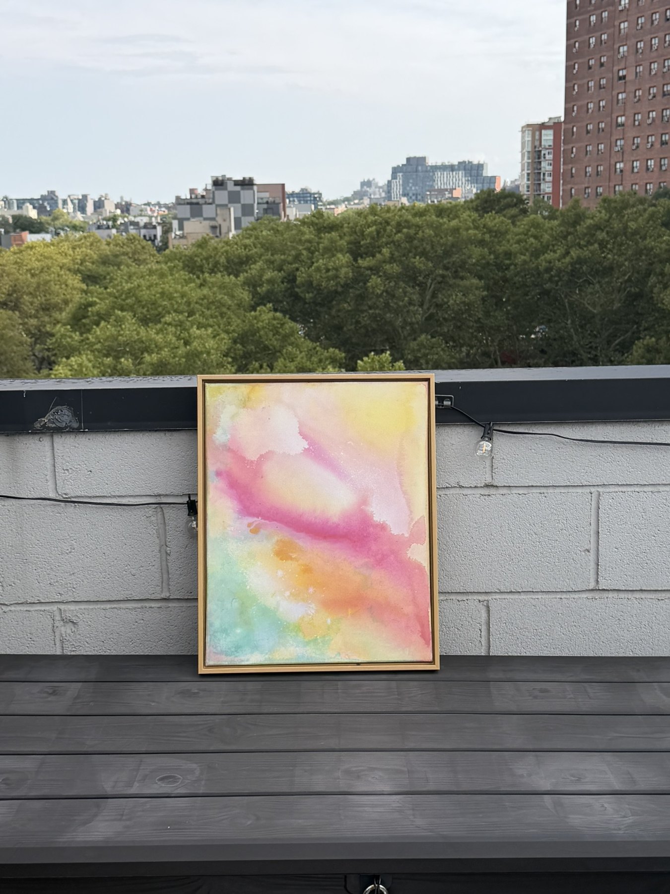 A small framed canvas in soft yellow, pink, orange and mint washes, propped on a rooftop ledge with trees and buildings behind.