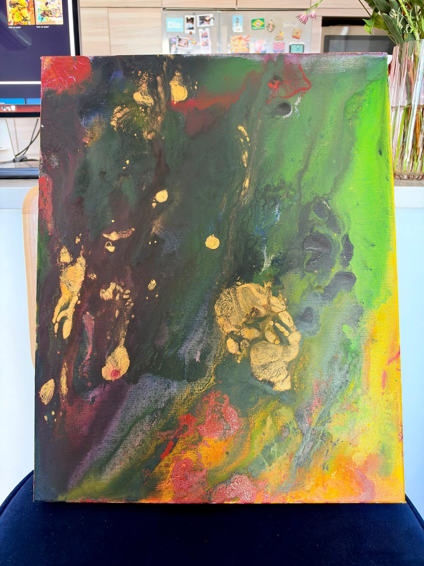 A small canvas in deep green and black washes with gold pours and red, breaking into bright green and yellow on the right edge, standing on a chair indoors.